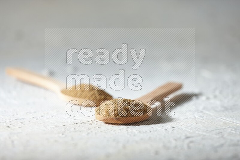 2 Wooden spoons full of cumin powder on a textured white flooring