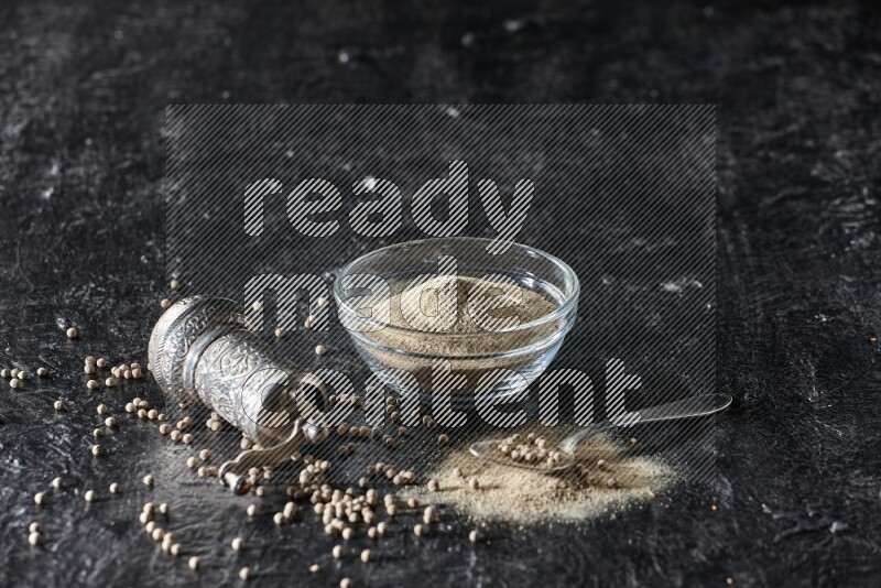 A glass bowl full of white pepper powder with pepper beads, a metal grinder and a metal spoon on textured black flooring