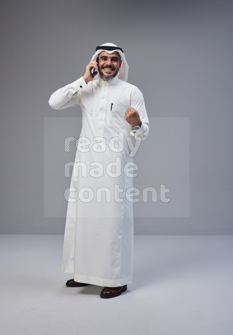 Saudi man Wearing Thob and white Shomag standing talking on phone on Gray background