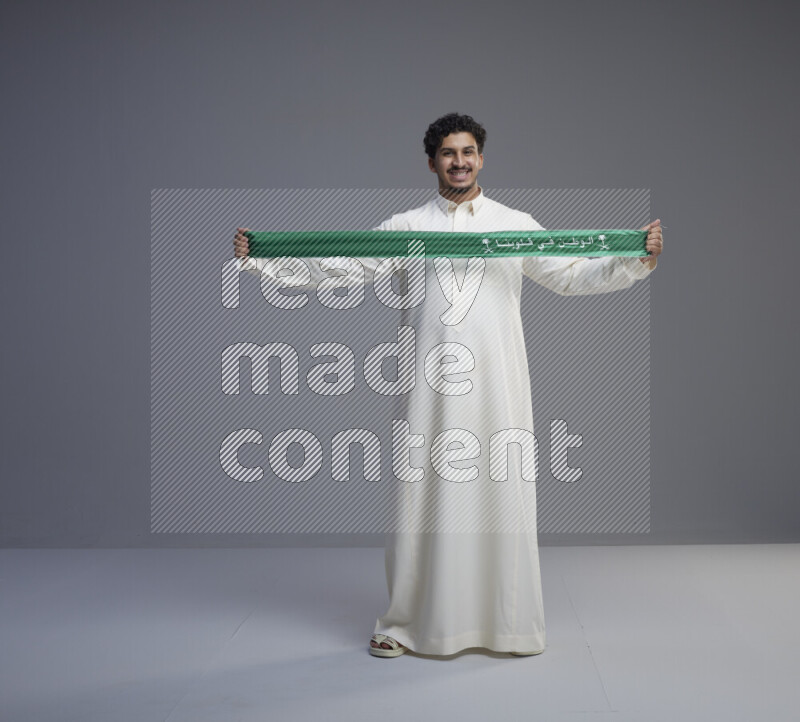 A Saudi man standing wearing thob holding Saudi flag scarf on gray background