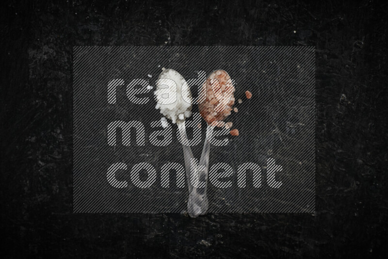 2 metal spoons filled with white salt and pink himalayan salt on black background