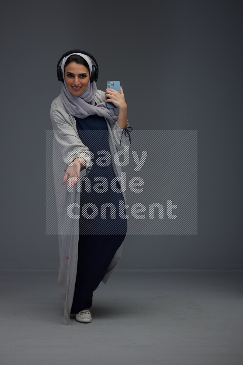 A Saudi woman wearing a light gray Abaya and head scarf standing and listening to music on a grey background