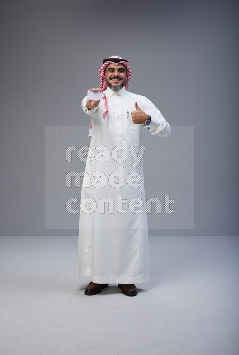 Saudi man Wearing Thob and red Shomag standing holding ATM card on Gray background