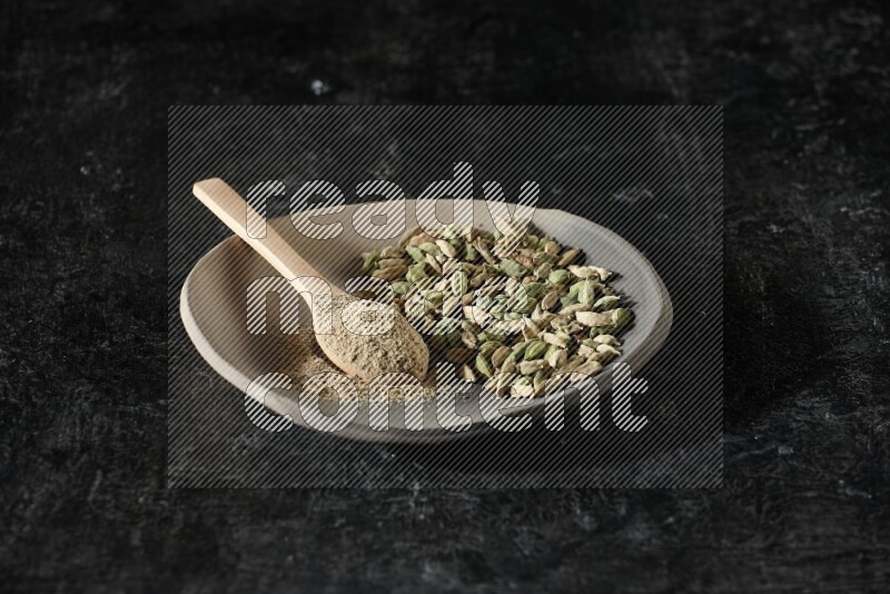 A plate filled with cardamom seeds and a wooden spoon full of cardamom powder on a textured black flooring