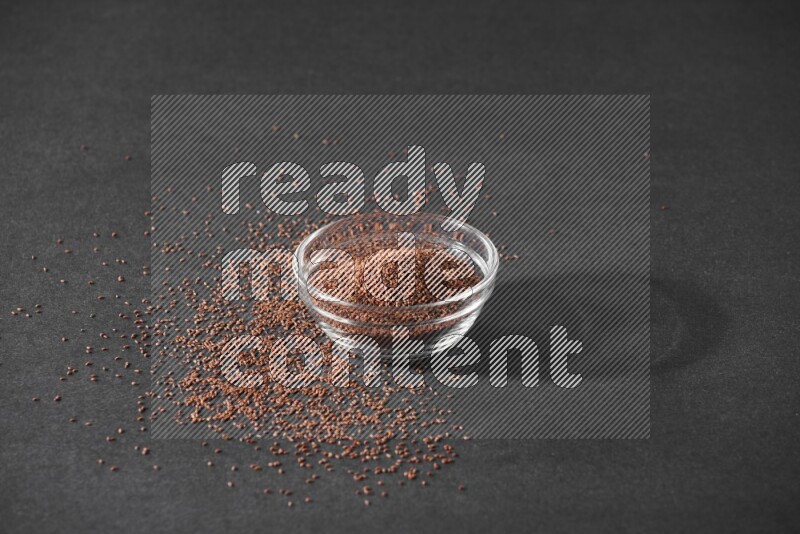 A glass bowl full of garden cress seeds surrounded by seeds on a black flooring