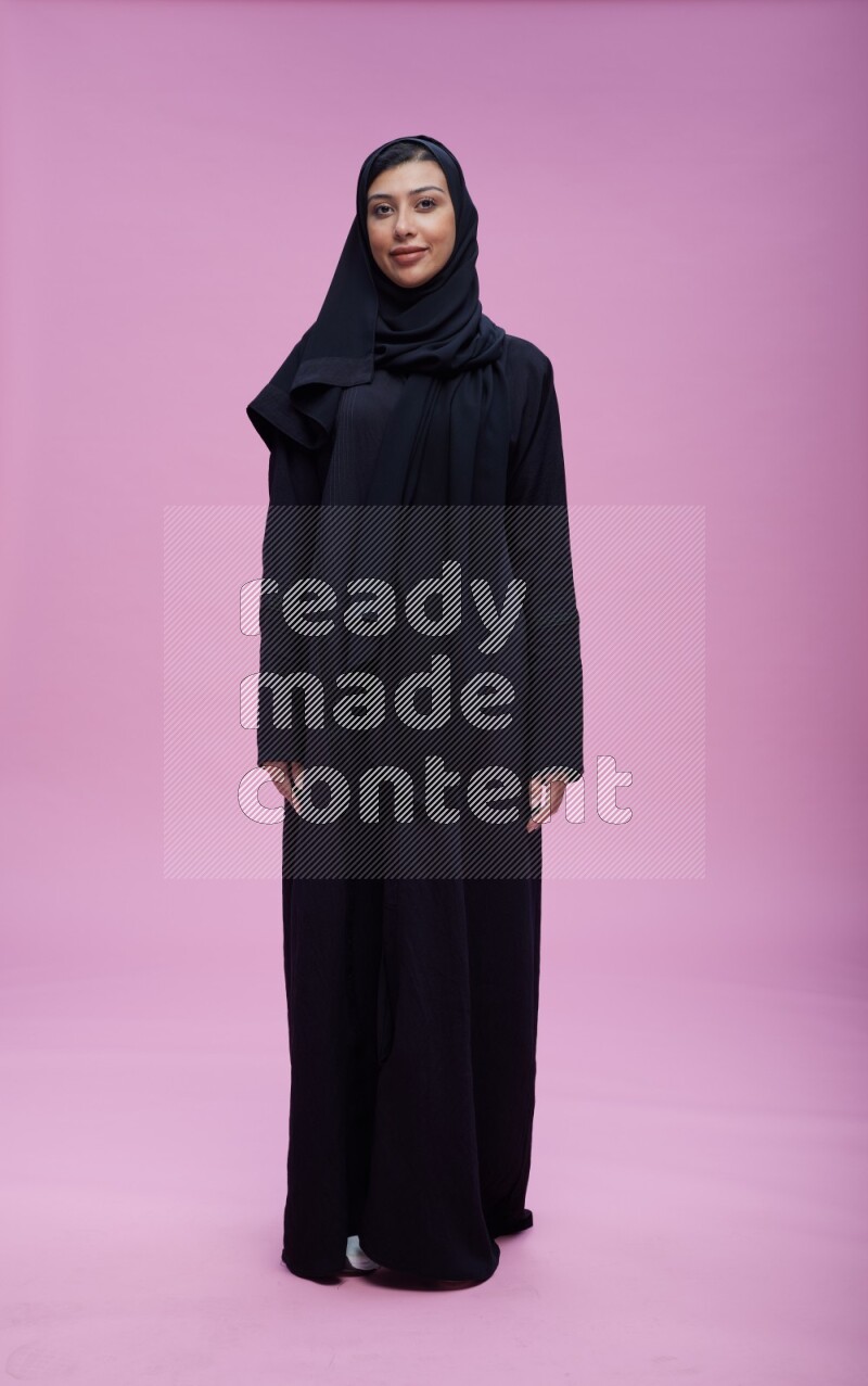 Saudi woman wearing Abaya standing interacting with the camera on pink background