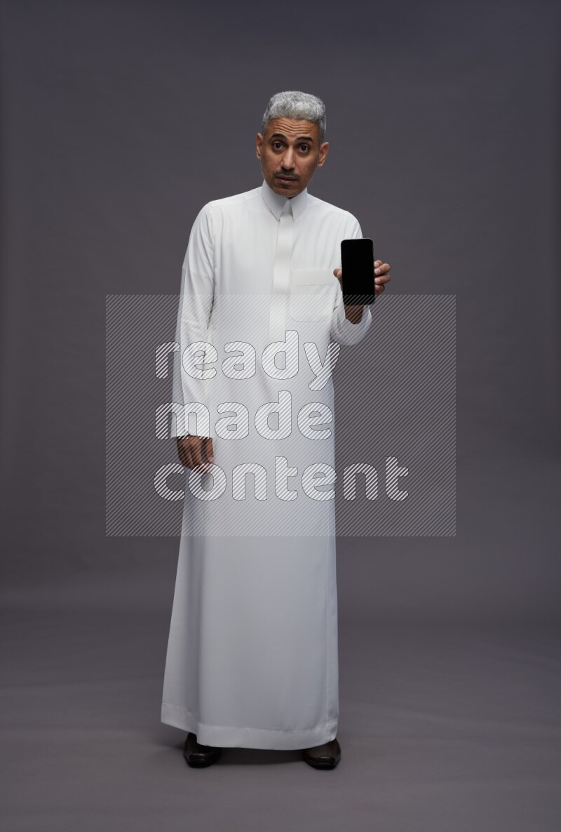 Saudi man wearing thob standing showing phone to camera on gray background