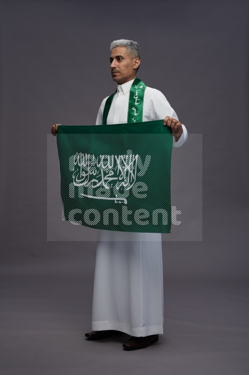 Saudi man wearing thob standing holding Saudi flag on gray background