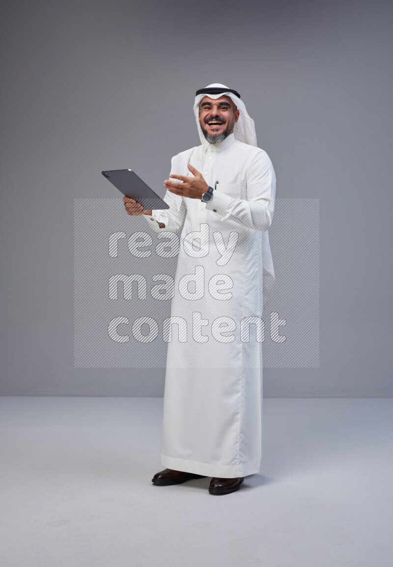 Saudi man Wearing Thob and white Shomag standing working on tablet on Gray background