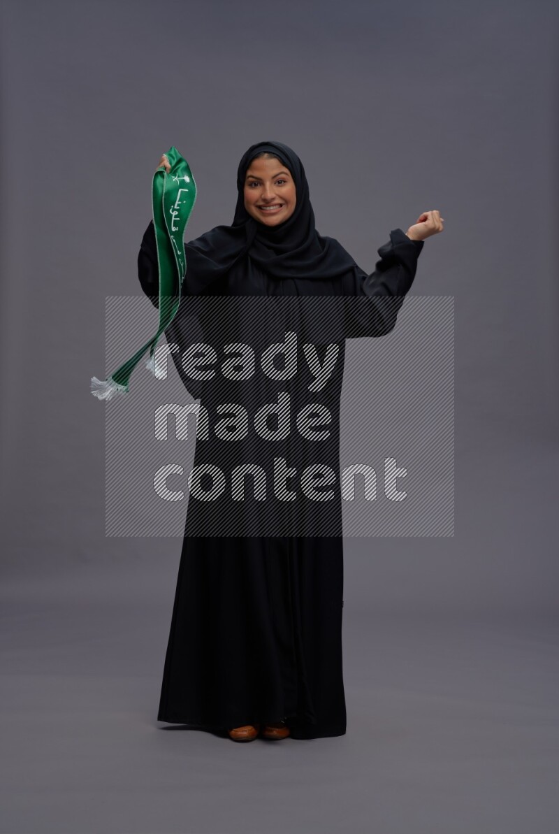 Saudi woman wearing Abaya standing holding Saudi flag on gray background