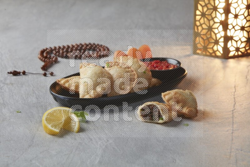 Five fried sambosas in an oval shaped black plate, beside a cut meat sambosa, a brown misbaha and a golden lantern on a gray background