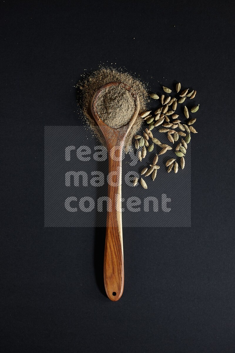 A wooden ladle full of cardamom powder and cardamom seeds beside it on black flooring