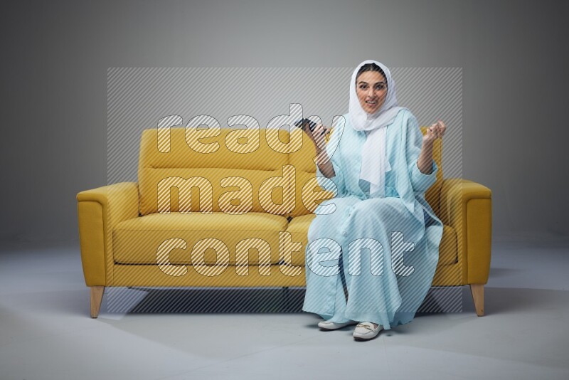 A Saudi woman wearing a light blue Abaya and white head scarf sitting on a yellow sofa and using her phone eye level on a grey background