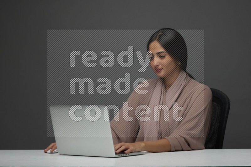 A Saudi woman Sitting on her desk on a Gray Background wearing Brown Abaya