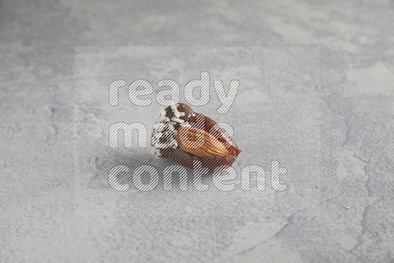 Almond stuffed date covered with dark chocolate and coconut on a light grey background