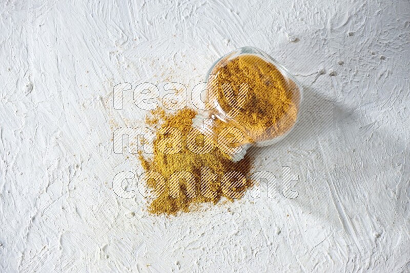 A flipped glass spice jar full of turmeric powder and powder fell out it on textured white flooring