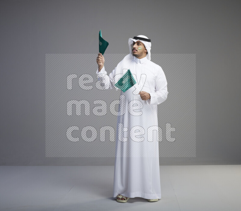 A Saudi man standing wearing thob and white shomag with face painting raising small saudi flag on gray background