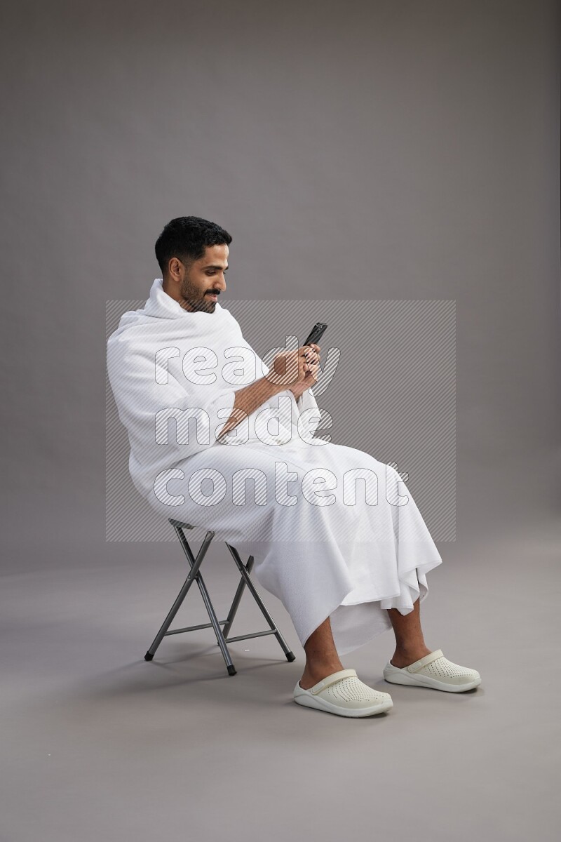 A man wearing Ehram sitting on chair texting on phone on gray background