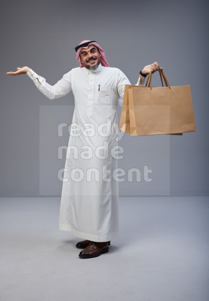 Saudi man Wearing Thob and red Shomag standing holding shopping bag on Gray background