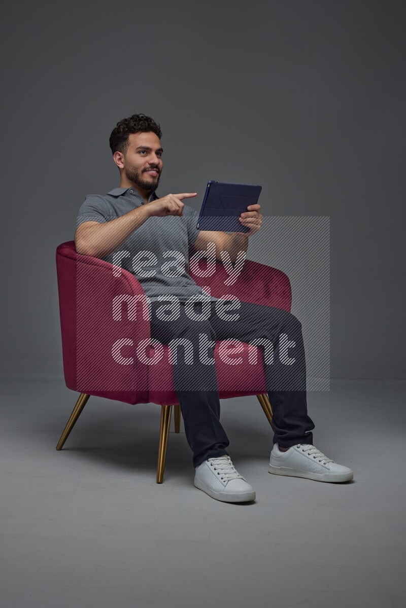A man wearing casual and using his tablet while sitting on a burgundy chair eye level on a gray background