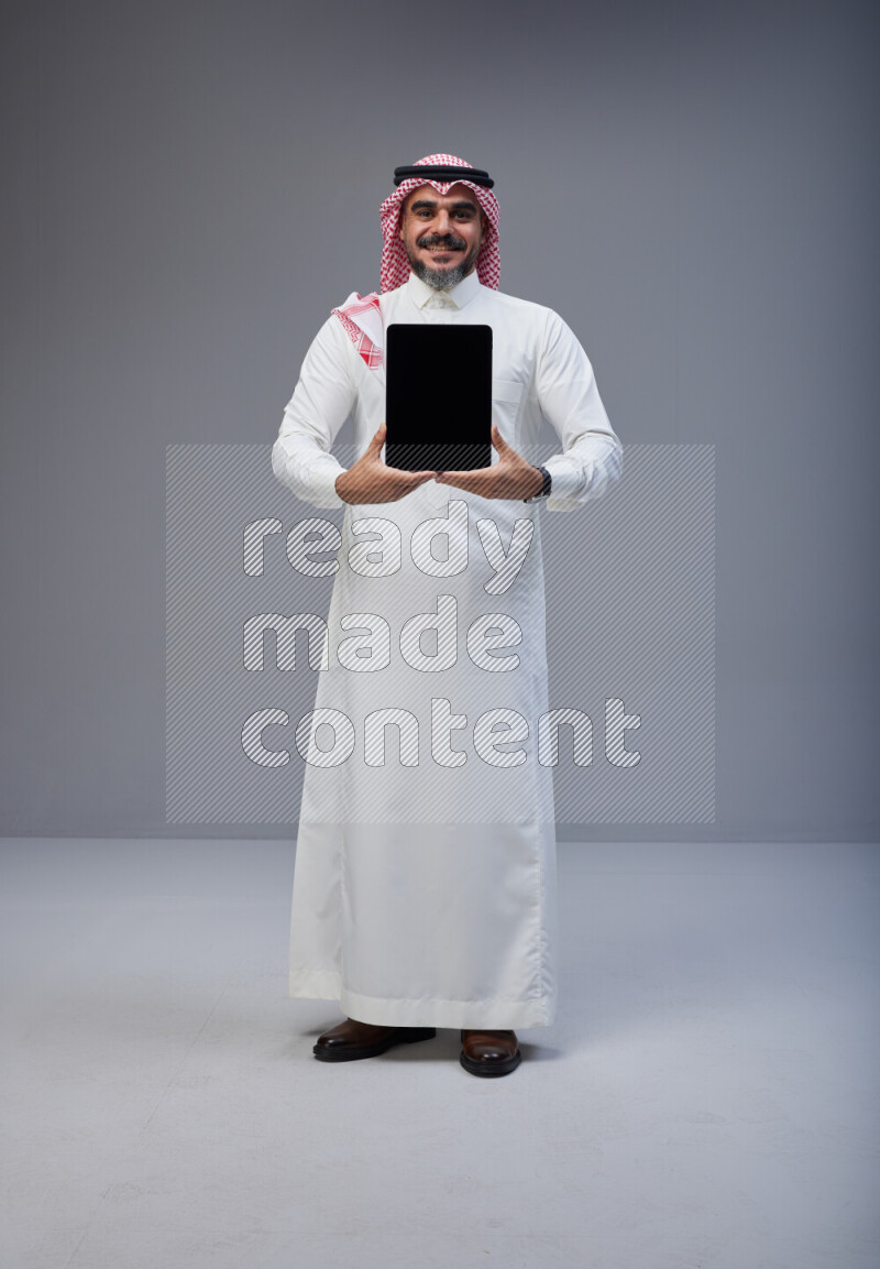 Saudi man Wearing Thob and red Shomag standing showing tablet to camera on Gray background