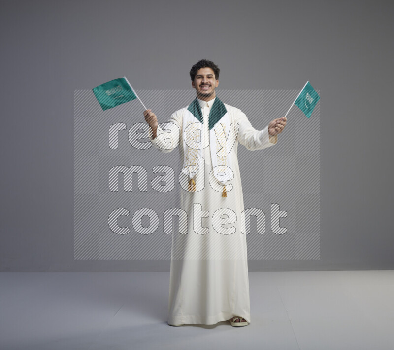 A Saudi man standing wearing thob and saudi flag scarf and holding small saudi flag on gray background