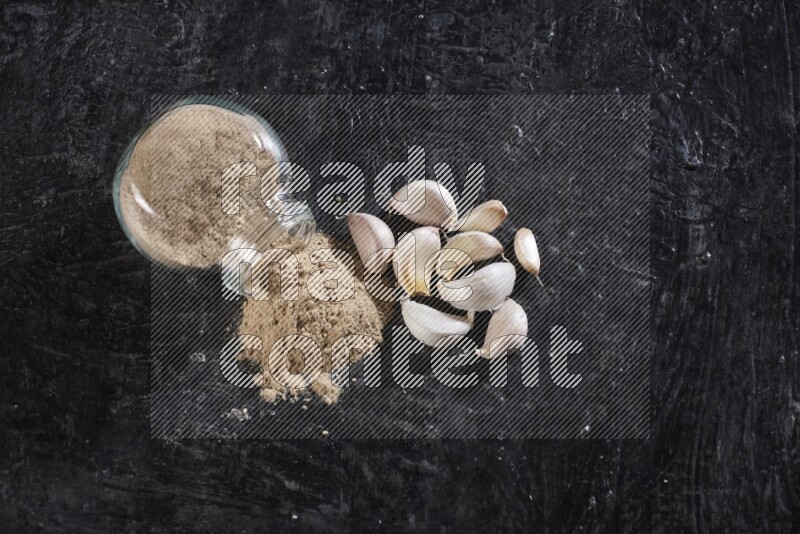 A glass spice jar full of garlic powder flipped over with the powder came out on a textured black flooring