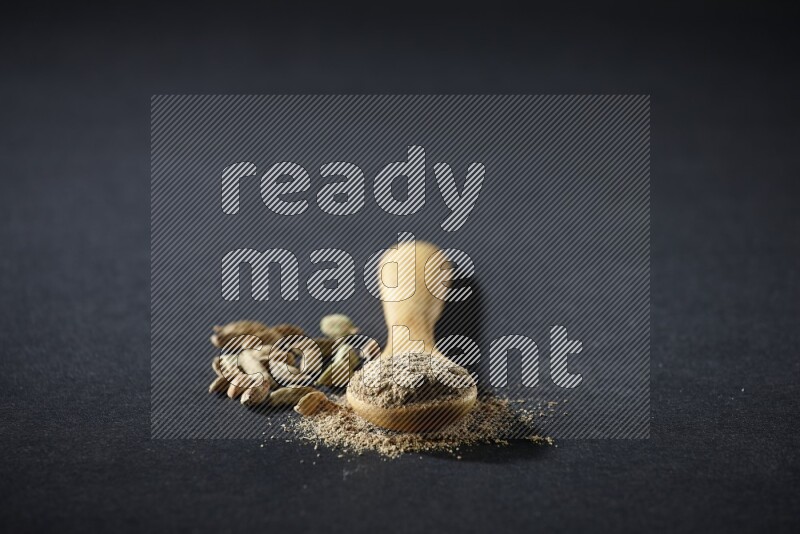 A wooden spoon full of cardamom powder and cardamom seeds beside it on black flooring