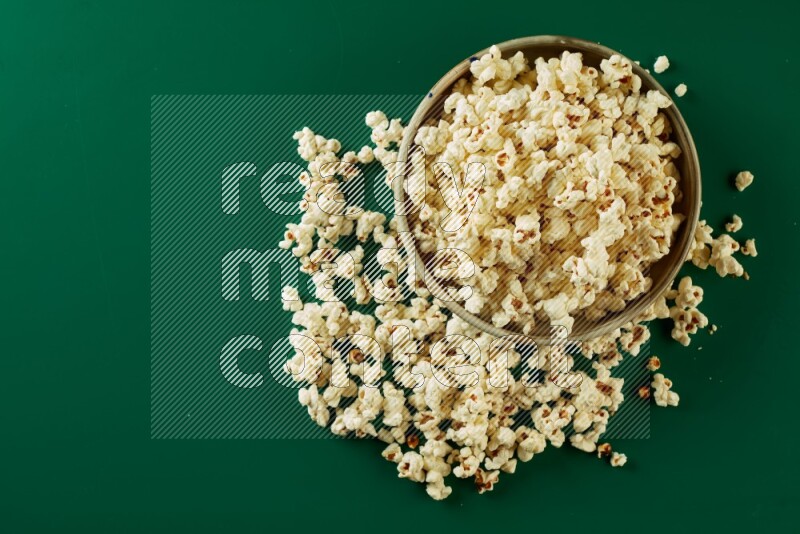 A beige ceramic bowl full of popcorn with popcorn beside it on a green background in a top view shot