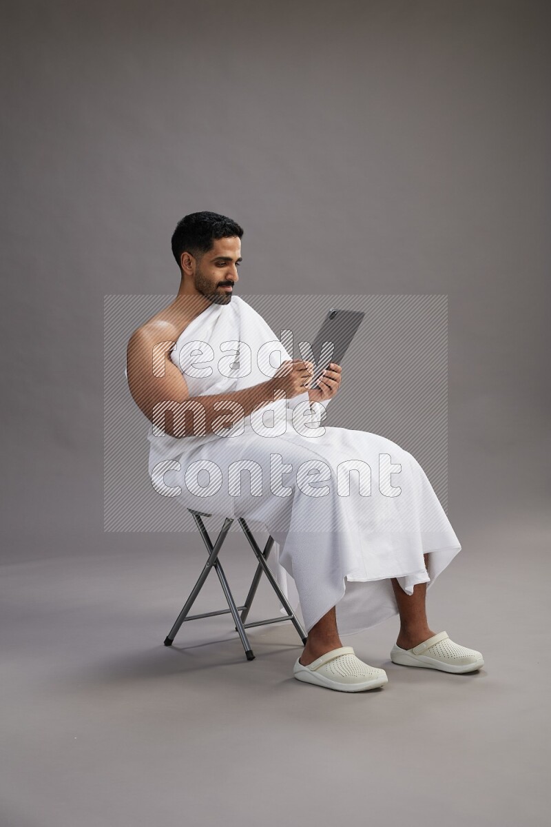 A man wearing Ehram sitting on chair holding a tablet on gray background