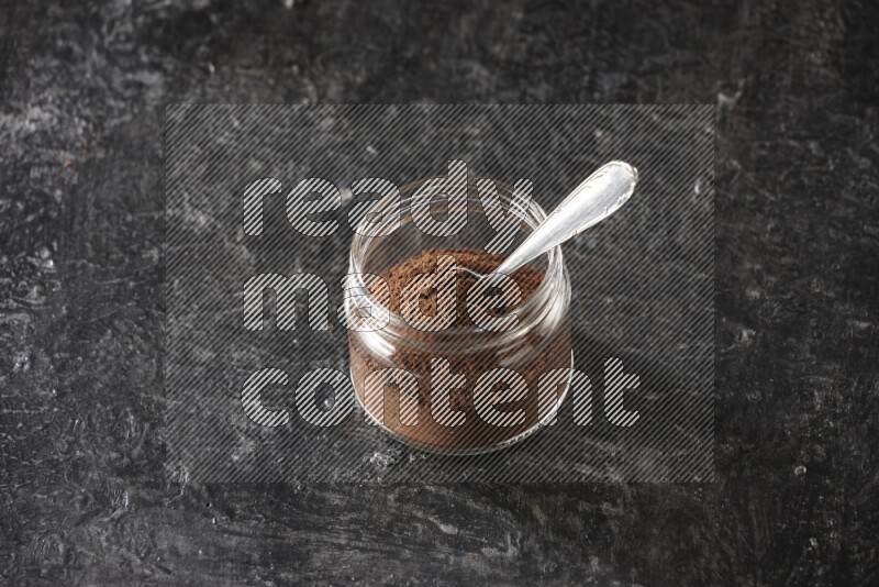 A glass jar full of cloves powder with a metal spoon on a textured black flooring