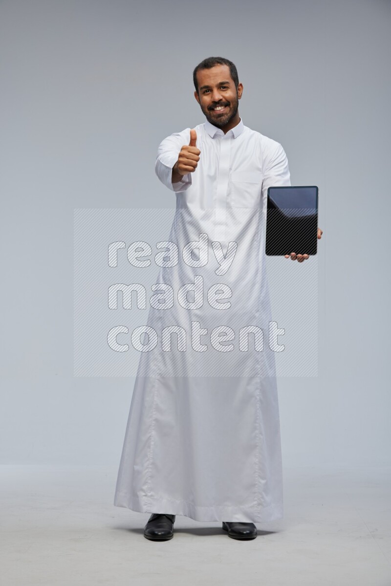 Saudi man wearing Thob standing showing tablet to camera on Gray background