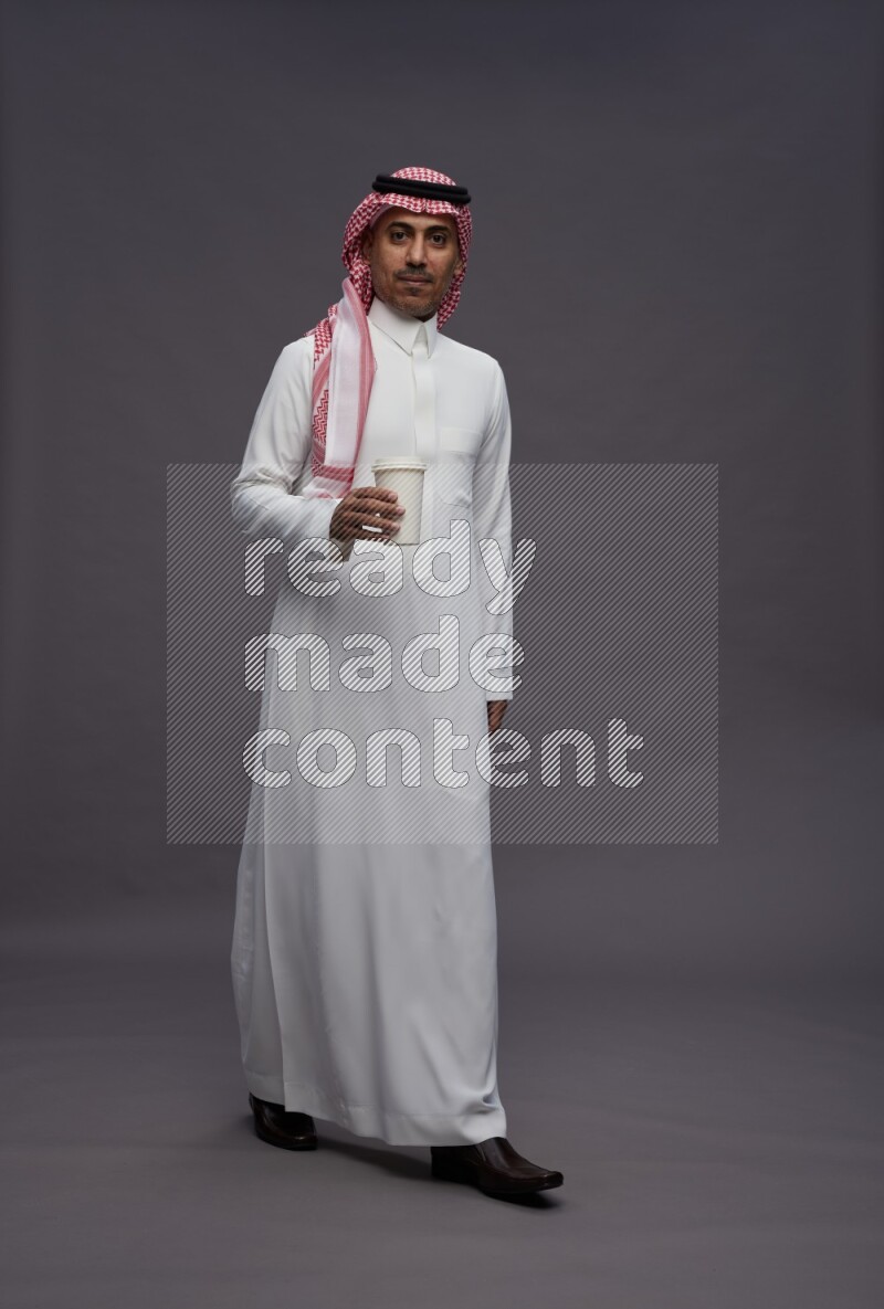 Saudi man wearing thob and shomag standing holding paper cup on gray background