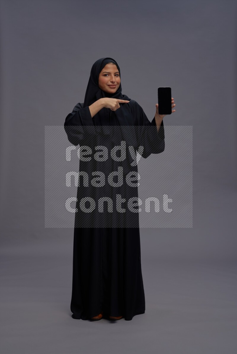 Saudi woman wearing Abaya standing showing phone to camera on gray background