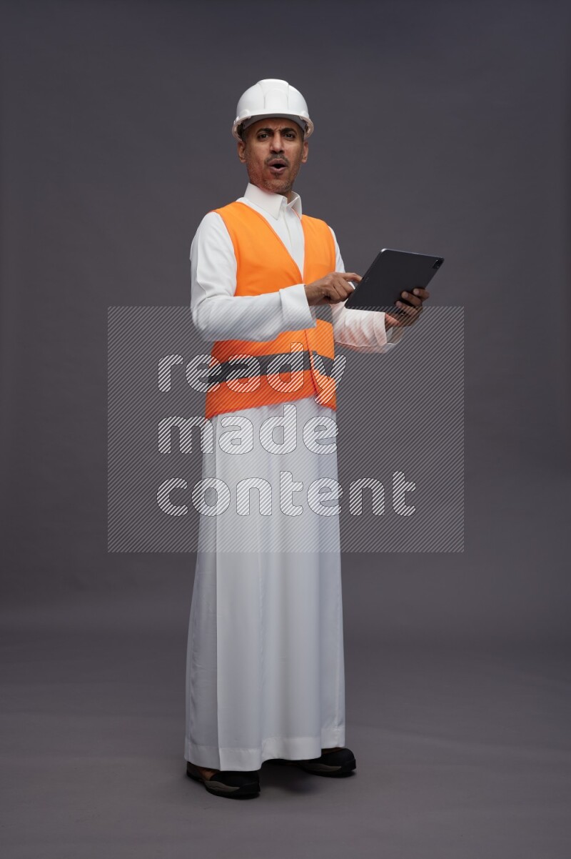 Saudi man wearing thob with engineer vest standing working on tablet on gray background