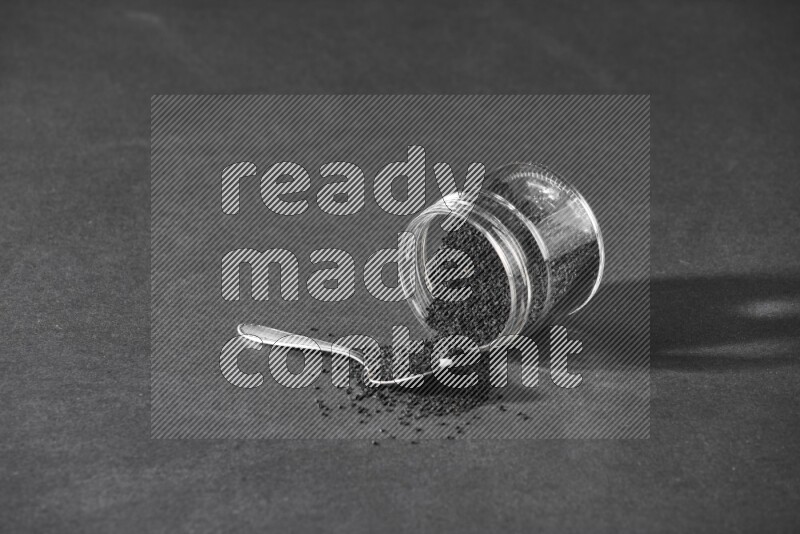 وعاء زجاجي مقلوب ممتلئ ببذور حبة البركة والبذور متناثرة بجانبه مع ملعقة معدنية ممتلئة بالبذور على أرضية سوداء