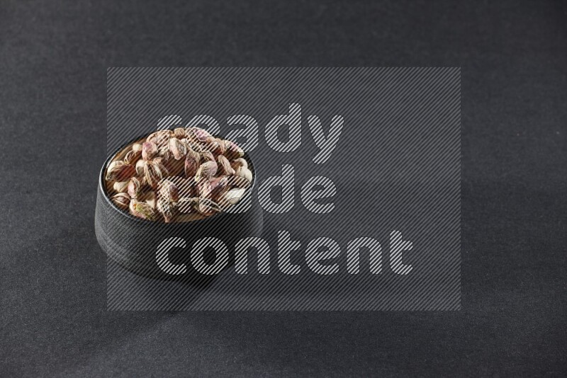 A black pottery bowl full of peeled pistachios on a black background in different angles