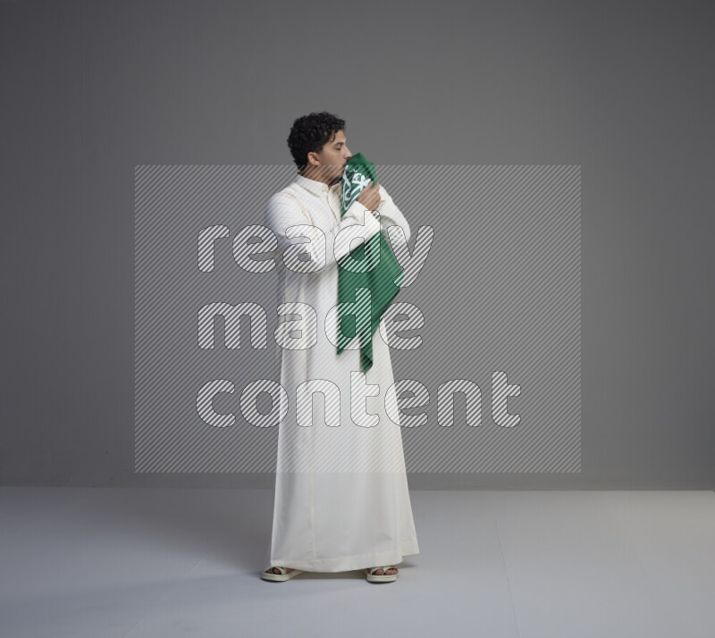 A Saudi man standing wearing thob kissing Saudi flag on gray background