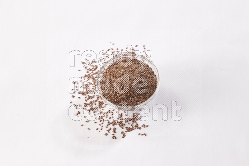 A glass bowl full of flax seeds surrounded by flax seeds on a white flooring