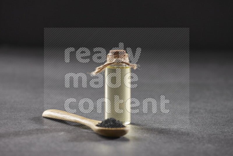 A wooden spoon full of black seeds and a glass bottle of black seeds oil on a black flooring