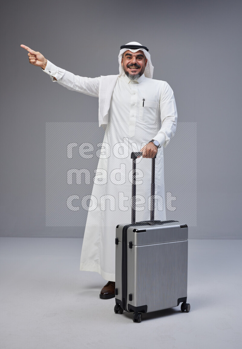 Saudi man wearing Thob and white Shomag standing holding Travel bag on Gray background