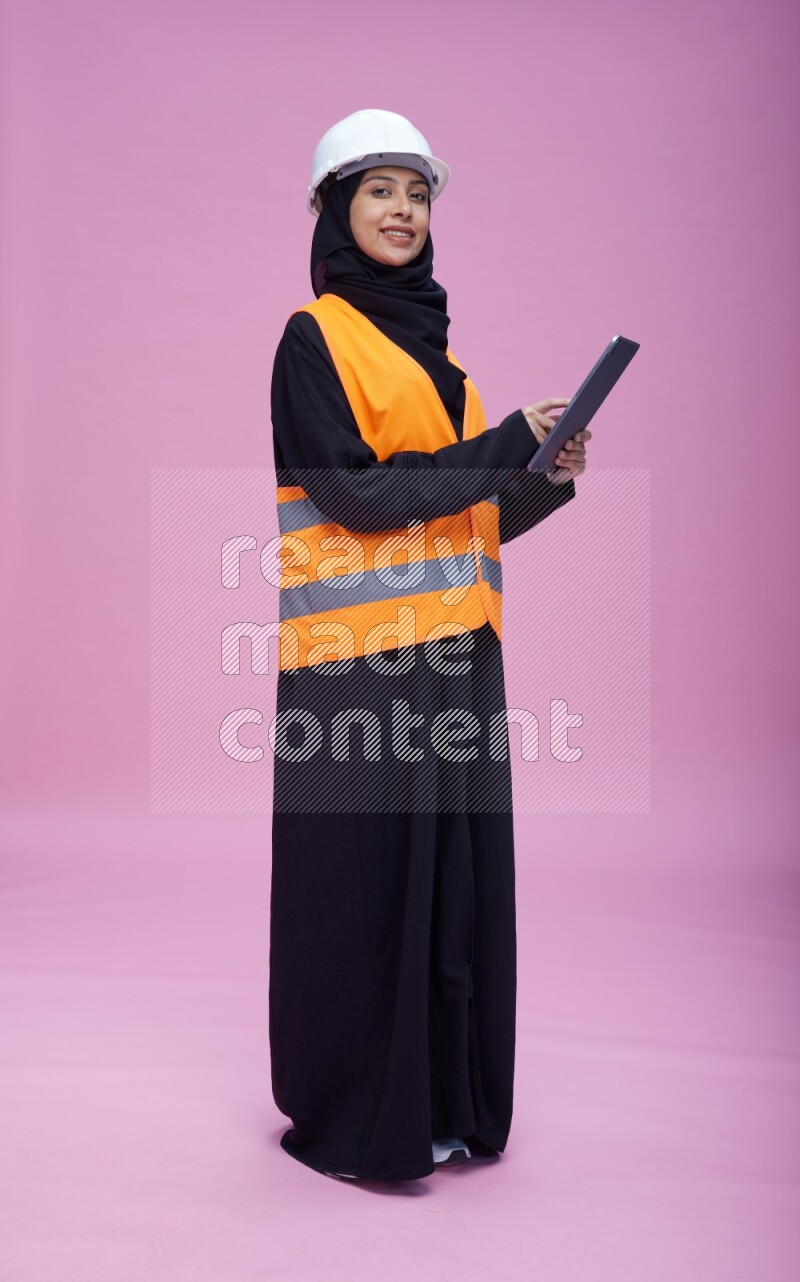 Saudi woman wearing Abaya with engineer vest and helmet standing working on tablet on pink background