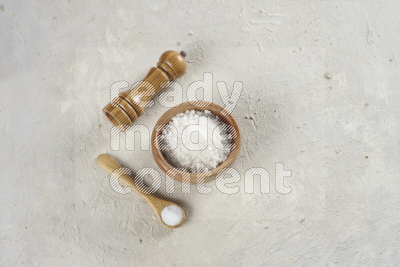 A wooden bowl and spoon filled with white sea salt and wooden grinder beside them on white background