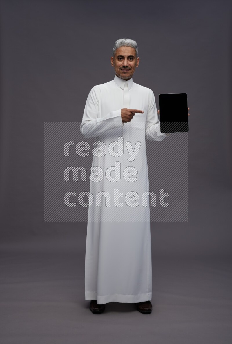 Saudi man wearing thob standing showing tablet to camera on gray background