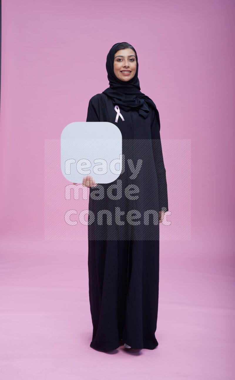 Saudi woman wearing pink ribbon on Abaya standing holding social media signs on pink background