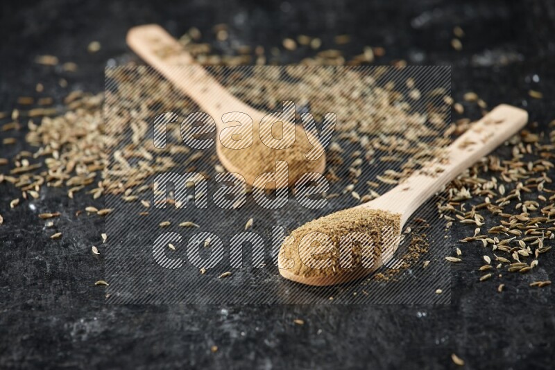 2 wooden spoons full of cumin powder with spreaded seeds on textured black flooring