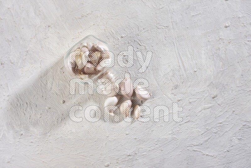 A glass spice jar full of garlic cloves flipped over with the cloves spilling out on a textured white flooring