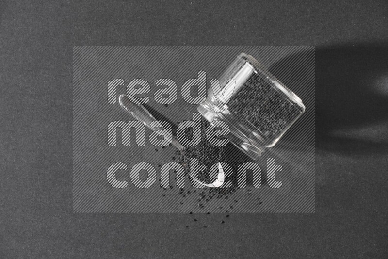 A flipped glass jar full of black seeds and seeds spread out with a metal spoon full of the seeds on a black flooring