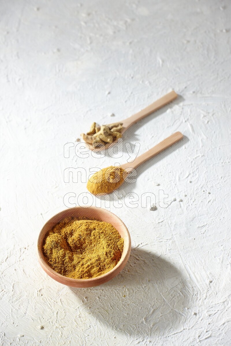 A wooden bowl full of turmeric powder and 2 wooden spoons full of dried turmeric whole finger and turmeric powder on textured white flooring