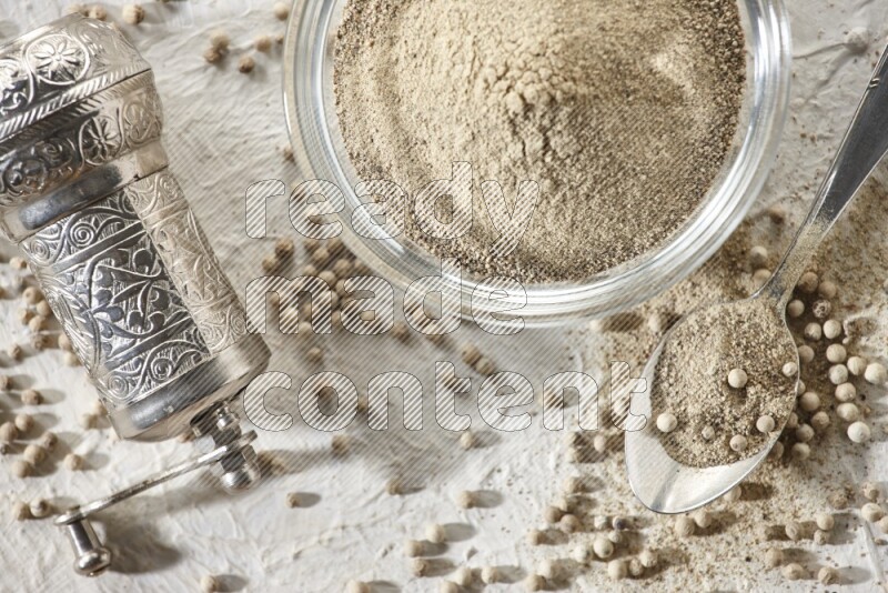 A glass bowl full of white pepper powder with white pepper beads and a metal grinder on textured white flooring